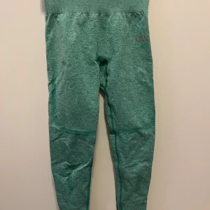 Gröna leggings från Gymshark - Snygga gröna leggings från Gymshark med hög midja och ribbade detaljer. Perfekta för träning med en tight passform som ger bra stöd. Materialet är stretchigt och bekvämt, vilket gör dem idealiska för intensiva träningspass.