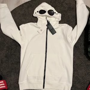 Vit hoodie från C.P. Company - Säljer en stilren vit hoodie från C.P. Company med dragkedja och unika glasögon i huvan. Perfekt för dig som vill ha något annorlunda och trendigt. Den har en broderad logga på bröstet och är tillverkad i mjukt material för extra komfort. Koftan är helt ny. Priset går att diskuteras för mer info eller fler bilder så är det bara att skriva. VIKTIGT TRÖJAN PASSAR STORLEK (S) SITTER SOM TAJT (M) !SKCIKAS SAMMA DAG ELLER DAGEN EFTER!
