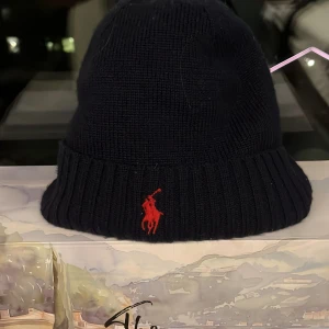 Svart mössa från Polo Ralph Lauren - Snygg mörkblå mössa från Polo Ralph Lauren i merinoull. Den har en ribbad kant och ett rött broderat emblem framtill. Perfekt för att hålla värmen med stil.