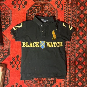 Svart pikétröja från Ralph Lauren - Säljer en svart pikétröja från Ralph Lauren med gul text 'BLACK WATCH' och nummerdetaljer på ärmarna och ryggen. Tröjan har en klassisk krage och korta ärmar. Perfekt för en sportig look!