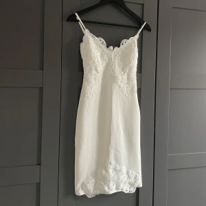 Vit spetsklänning  - Vit figurnära klänning med spetsdetaljer. Klänningen har smala axelband och en dragkedja baktill. I storlek EU32, men väldigt stretchig i materialet.