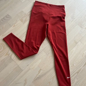 Roströda leggings från Nike - Snygga röda leggings från Nike med hög midja och en liten vit logga vid ankeln. Perfekta för träning eller en avslappnad dag. Tillverkade i ett stretchigt syntetmaterial för bästa komfort och passform.