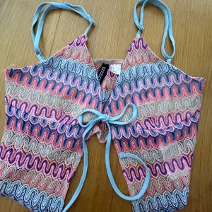 Flerfärgad topp från Divided - Säljer en färgglad strandtopp från divided i missoni liknanade mönster. Toppen har smala axelband och en knytning framtill. Perfekt inför sommaren. Är något liten i storleken.