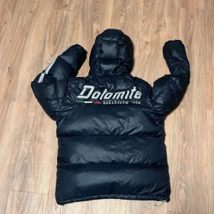 Blå dunjacka från Dolomite - Säljer en svart dunjacka från Dolomite med huva och dragkedja. Jackan har en stor logga på ryggen och mindre logga på bröstet. Perfekt för kalla dagar med sin pufferdesign och bekväma passform.
