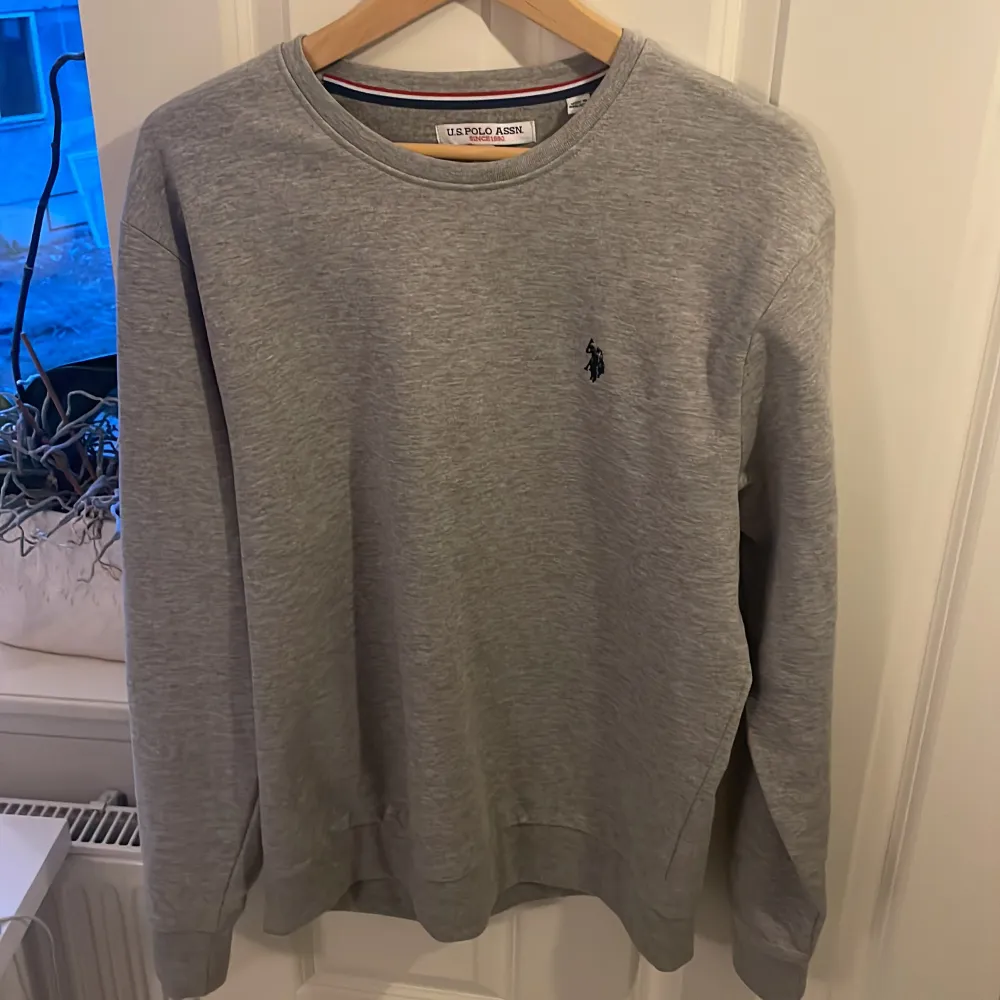 Säljer en stilren grå tröja från U.S. Polo Assn. med rund hals och det klassiska logotypbroderiet på bröstet. Tröjan har långa ärmar och är perfekt för en avslappnad look.. Neuletakit & Villapaidat.