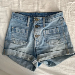 Blå jeansshorts från Levi's - Säljer ett par snygga blå jeansshorts från Levi's med hög midja och uppvikta ben. Shortsen har knappar framtill och två fickor både fram och bak. 