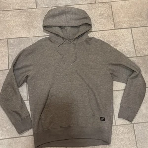 Grå hoodie från Jack & Jones - Säljer en stilren grå hoodie från Jack & Jones. Den har en klassisk design med huva och dragsko samt en praktisk magficka. Perfekt för en avslappnad stil.