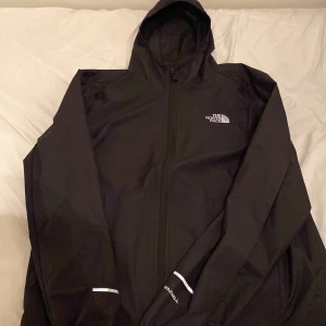 Svart vindjacka från The North Face - Säljer en svart vindjacka från The North Face. Jackan har en huva och dragkedja framtill. Perfekt för blåsiga dagar och har en stilren design med The North Face-logga på bröstet. Kan gå ner i pris vid snabb affär