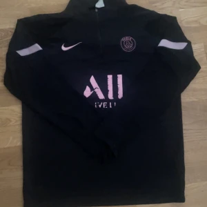 Svart långärmad tröja från Nike - Säljer en svart långärmad tröja från Nike med rosa detaljer och PSG-logga. Tröjan har en halv dragkedja framtill och texten 'Accor Live Limitless' på bröstet. Perfekt för fotbollsfans!