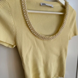 Gul ribbad topp med pärldetaljer från Zara S  - Säljer en söt gul ribbad topp från Zara med korta ärmar och en vacker pärlkrage. Perfekt för att ge en touch av elegans till din outfit. Toppen har en tight passform och är gjord i ett mjukt material. Storlek S använd vid enstaka tillfälle. 