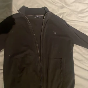 Svart ziptröja från Gant - Säljer en stilren svart ziptröja från Gant med dragkedja framtill. Jackan har en ribbad krage och är långärmad. Perfekt för en avslappnad look.