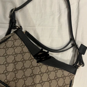 Mönstrad axelväska med svart läderdetalj - Snygg axelväska med klassiskt mönster och svarta läderdetaljer. Väskan har en justerbar axelrem och dragkedja upptill. Perfekt för att ge din outfit en stilfull touch.