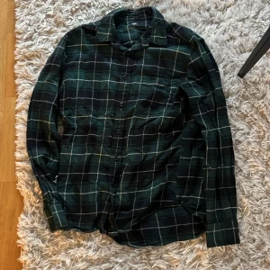 Grön och svart rutig skjorta Uniqlo - Snygg grön och svart rutig skjorta från Uniqlo med långa ärmar och klassisk krage. Perfekt för en avslappnad stil. Skjortan har knappar framtill och är tillverkad i ett mjukt material.