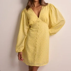 Gul klänning med v-ringning - Charmig gul klänning med v-ringning och långa ärmar. Den har ett diskret prickigt mönster och en omlott-design framtill. Perfekt för en solig dag!