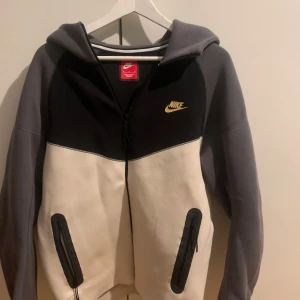 Nike tech  - Säljer en snygg huvtröja från Nike i grått och vitt med dragkedja framtill. Tröjan har en klassisk design med Nike-logga på bröstet och praktiska fickor med dragkedja. Perfekt för en avslappnad stil. Nyskick använd 1 gång, skick 10 av 10 pris kan diskuteras vid snabb afär 