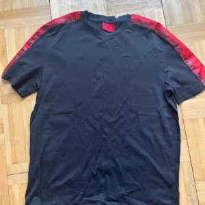 Svart t-shirt med röda detaljer från Hugo - Snygg svart t-shirt från Hugo med röda band och logga längs ärmarna. Perfekt för en stilren look med en touch av färg. T-shirten har en klassisk passform och är kortärmad.