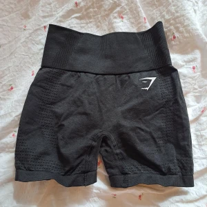 Svarta träningsshorts från Gymshark - Säljer ett par svarta träningsshorts från Gymshark med hög midja och ribbad detalj. Perfekta för gymmet eller löprundan. De är tillverkade i ett stretchigt syntetmaterial för optimal rörelsefrihet.