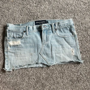 Ljusblå jeanskjol från Abercrombie - Trendig ljusblå jeanskjol från Abercrombie med slitna detaljer och rå kant. Perfekt för en avslappnad stil. Kjolen har klassiska fem fickor och bälteshällor.