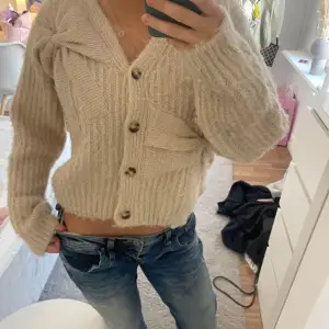 Mysig beige stickad kofta med stora knappar framtill. Koftan har en croppad passform och långa ärmar med ribbade muddar. Perfekt för kyliga dagar!