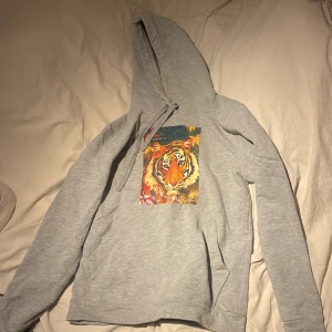 Grå hoodie med tigertryck - Säljer en grå hoodie med ett färgglatt tigertryck på framsidan. Tröjan har en klassisk huva med dragsko och långa ärmar. Perfekt för en avslappnad stil.