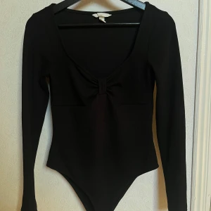 Svart långärmad bodysuit med rosett - Snygg svart långärmad bodysuit med en söt rosett framtill. Perfekt för en stilren look. Den har en djup v-ringning och är tillverkad i ett stretchigt material för en bekväm passform.