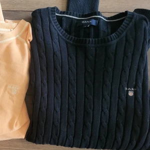 Svart kabelstickad tröja från GANT - Säljer en stilren svart kabelstickad tröja från GANT med rund halsringning. Tröjan har ett diskret broderat GANT-emblem på bröstet.