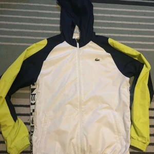 Vit och gul vindjacka från Lacoste - Säljer en snygg vit och gul vindjacka från Lacoste med dragkedja och huva. Jackan har långa ärmar och Lacoste-logga på bröstet samt längs sidorna. Perfekt för vår och höst!