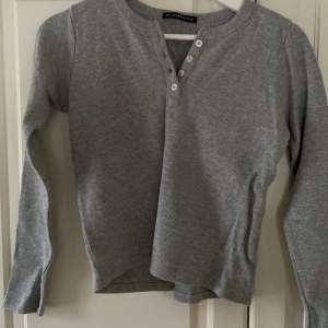 Säljer en grå ribbad tröja från Brandy Melville med knappar framtill. Tröjan har långa ärmar och en bekväm passform. Perfekt för en avslappnad stil.