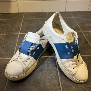 Valentino Garavani sneakers i vitt och blått - Snygga Valentino Garavani sneakers i vitt med blå detaljer. Skorna har en unik design med breda blå remmar över snörningen och rund tå. Perfekta för en stilren look. Lite öppna stygn längst fram bara men det går att fixas☺️