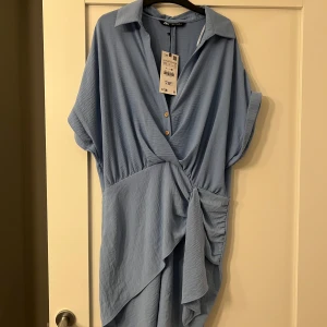 Blå omlottklänning från Zara - Säljer en snygg blå omlottklänning från Zara med knappar framtill och en elegant drapering. Klänningen har korta ärmar och en krage som ger en stilren look. Perfekt för en avslappnad men chic stil.