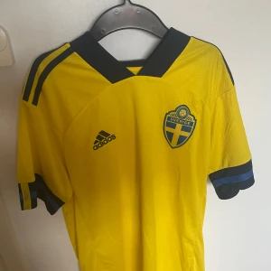 Gul fotbollströja från Adidas - Säljer en gul fotbollströja från Adidas med Sveriges landslagsemblem på bröstet. Tröjan har korta ärmar och mörkblå detaljer på krage och ärmar. Perfekt för fotbollsfans som vill visa sitt stöd för Sverige! 💛💙