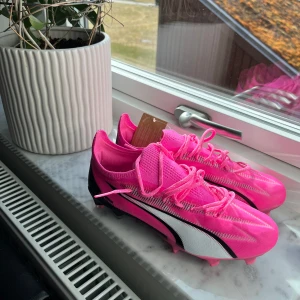 Rosa fotbollsskor från Puma - Säljer ett par snygga rosa fotbollsskor från Puma med vita och svarta detaljer. Skorna har snörning och är perfekta för konstgräs. De är i storlek dam 37,5 och har en modern design som sticker ut på planen. Säljer tyvärr pga för stora:(  nypris  1499 säljer för 1100