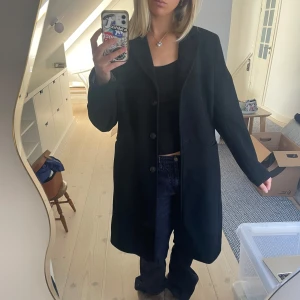 Svart kappa New House - Snygg svart kappa från New House med knappar framtill. Perfekt för kyligare dagar och ger en stilren look. Vadderad och varm. 