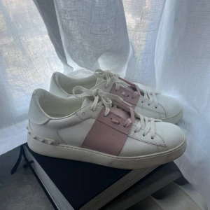 Vita Valentino sneakers med rosa detalj  - Snygga vita sneakers med en bred rosa rem över snörningen. Skorna har en rund tå och vita snören. Perfekta för en stilren look med en touch av färg.