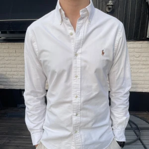 Vit skjorta från Ralph Lauren - Säljer en klassisk vit skjorta från Ralph Lauren med broderad logga på bröstet. Skjortan har långa ärmar och knappar framtill. Perfekt för en stilren look. Storleken är slim fit M och modellen är 185cm lång och väger 82kg.