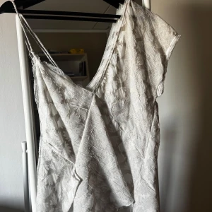 Vit klänning med blommigt mönster - Säljer en vacker vit klänning med blommigt mönster och smala axelband. Klänningen har en omlott-design och är perfekt för sommarens alla tillfällen. Den har en luftig och elegant stil som passar både till vardag och fest.