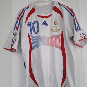 Vit fotbollströja från Adidas - Säljer en vit fotbollströja från Adidas med blå och röda detaljer. Tröjan har korta ärmar och nummer 10 på framsidan och baksidan. Den är I storlek medium. Pris kan diskuteras!