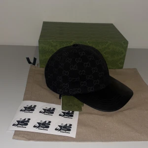 Svart keps från Gucci - Snygg svart keps från Gucci med det ikoniska GG-mönstret. Kepsen har en justerbar rem baktill och en böjd skärm i läder. Perfekt för att ge din outfit en lyxig touch. Finns i S M L