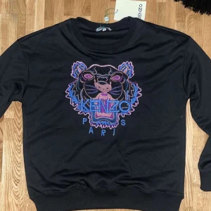 Svart Kenzo tröja  - Snygg svart Kenzo med ett iögonfallande broderi av en tiger i lila och blått på framsidan. Tröjan har rund hals och långa ärmar, perfekt för en stilren look. Den är storlek M men skulle säga den passar mer som S