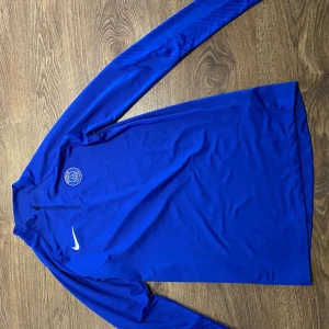 Blå långärmad tröja från Nike - Säljer en blå långärmad tröja från Nike med Paris Saint-Germain-logga på bröstet. Tröjan har en dragkedja vid halsen och är tillverkad i Dri-FIT-material för optimal komfort. Perfekt för fotbollsträning eller vardagsbruk.