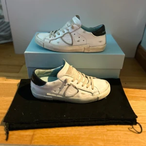 Vita Philippe Model - Snygga vita sneakers från Philippe Model med svarta detaljer på hälen. Skorna har en klassisk design med snörning och perforerade detaljer på sidorna. 9/10 bra skick. 🔥
