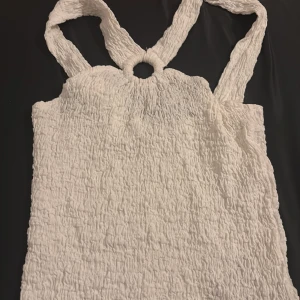 Vit ärmlös topp med ringdetalj - Säljer en vit ärmlös topp från Zara med en unik ringdetalj vid halsen. Toppen har en skrynklig textur som ger en cool look. Perfekt för varma dagar eller som ett lager under en jacka.