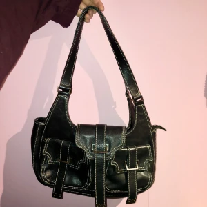 2000s axelväska - Snygg svart axelväska i skinn med kontrasterande sömmar och två framfickor med spännen. Väskan har ett blommigt foder i beige och lila samt en dragkedja för säker förvaring. Rymlig.