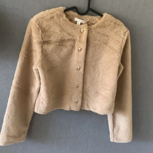Beige päls tröja från H&M - Säljer en supermjuk beige pälsjacka från H&M. Jackan har en rund halsringning och knäpps med eleganta knappar framtill. Aldrig använd