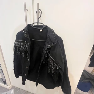Svart jacka med fransar och nitar - Snygg svart jacka med fransar och nitar som ger en cool look. Jackan har knappar framtill och långa ärmar. Perfekt för att sticka ut i mängden!