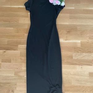 Svart klänning med blommig detalj - Elegant svart klänning med korta ärmar och en vacker blommig detalj vid axeln. Perfekt för en stilren look med en touch av färg.