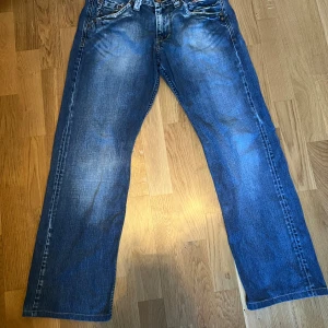 Blå jeans med broderade detaljer - Säljer ett par blå jeans med en avslappnad passform och broderade detaljer på bakfickorna. Jeansen har en klassisk femficksdesign och en lätt tvättad färg