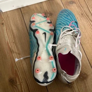 Nike fotbollsskor - Snygga fotbollsskor från Nike med ett unikt blått och vitt mönster. Skorna har snörning och en rund tå, med rosa detaljer som ger en extra touch. Perfekta för fotbollsplanen med sina greppvänliga dobbar.