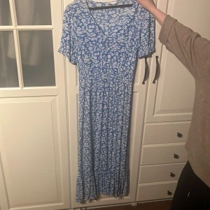 Blå blommig klänning från only  - Säljer en söt blå klänning med vita blommor. Klänningen har korta ärmar och en v-ringad hals. Perfekt för sommarens alla tillfällen. Den har en avslappnad passform och når ner till anklarna storlek M, köpt på only.