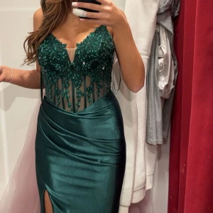 Grön långklänning med spetsdetaljer - Elegant grön långklänning med spetsdetaljer och öppen rygg. Klänningen har en v-ringad topp med pärlor och en slits framtill. Perfekt för festliga tillfällen.